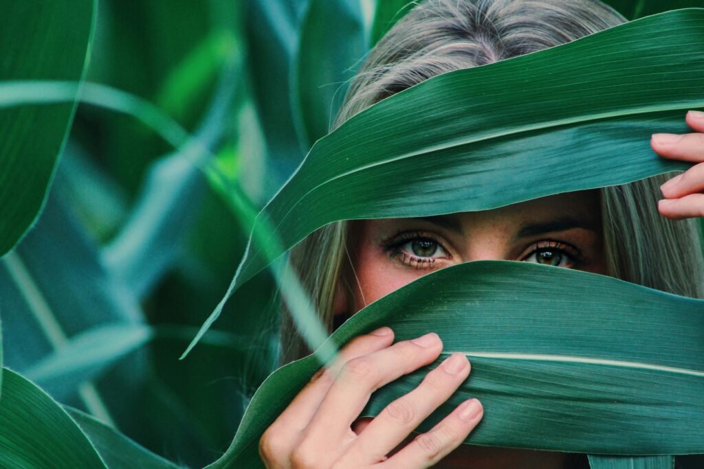 Close-up portrait of a young woman peeking through lush green leaves, in an outdoors setting.